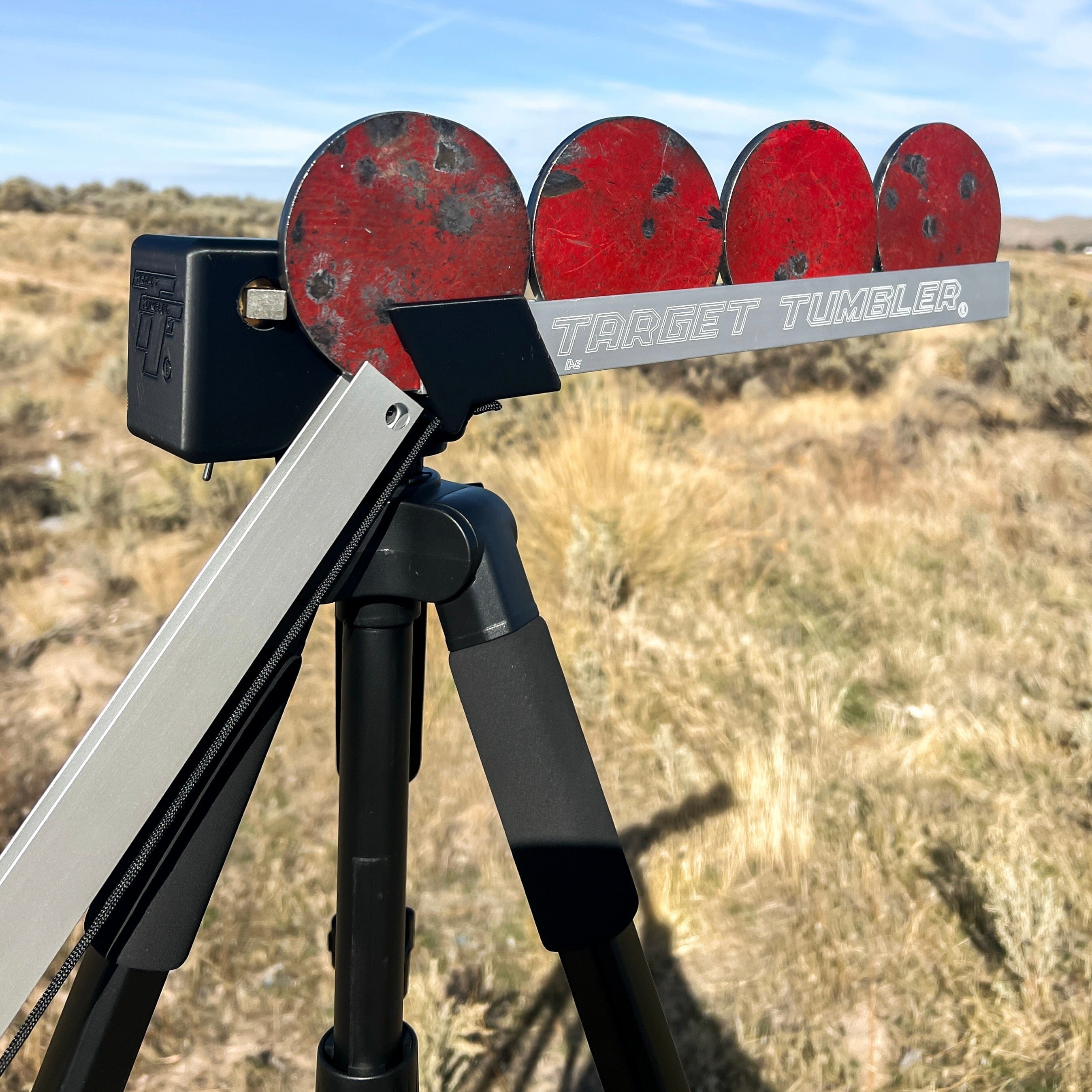 Red metal object with 'Target Tumbler' branding on a tripod in an outdoor setting.